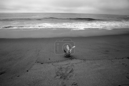 Foto de Cesta de Pascua en la arena junto al océano. Una cesta de Pascua con hierba falsa verde, un conejo de chocolate, huevos de plástico y orejas de conejo - Imagen libre de derechos