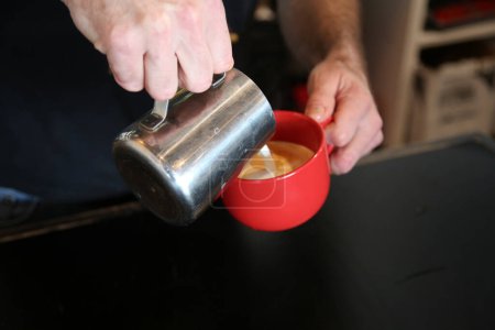 Téléchargez les photos : Latte versé par un avocat dans un café. Café tasse latte art. Une tasse de latte dans un café. Comment faire café latte art. Cappuccino avec mousse mousseuse dans une tasse rouge. - en image libre de droit