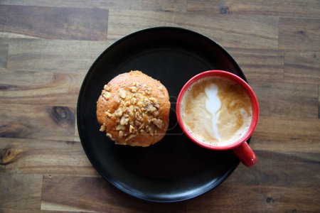 Foto de Latte con un Muffin de nuez de plátano. Café con una magdalena en un plato negro en una cafetería. Desayuno Muffin y Café con Latte Art en la espuma para el desayuno o un aperitivo en una cafetería. Desayuno. - Imagen libre de derechos