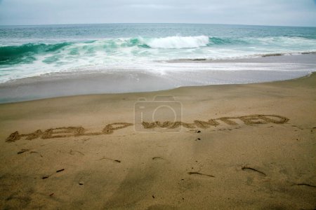 Téléchargez les photos : Aide voulait écrit dans le sable de plage avec l'océan comme arrière-plan. - en image libre de droit