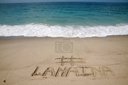 Téléchargez les photos : Le nom # LAHAINA écrit dans le sable sur la plage avec le fond de l'océan Pacifique - en image libre de droit