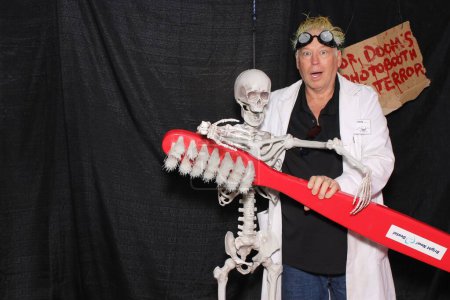 Photo for A man in a costume of an evil mad doctor poses with skeleton for Halloween while having his picture taken in a Photo Booth at a party. - Royalty Free Image