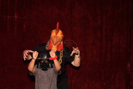 Téléchargez les photos : Une personne porte un masque de tête de poulet en caoutchouc tout en posant pour des photos dans une cabine photo avec un enfant. Les photomaton sont très amusants à toutes les fêtes et tous les événements. Tout le monde s'amuse avec des photos - en image libre de droit