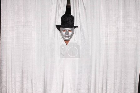 Foto de Un hombre con máscara de Halloween alcanza su punto máximo a través de cortinas blancas mientras está en una cabina de fotos en una boda o evento especial. - Imagen libre de derechos