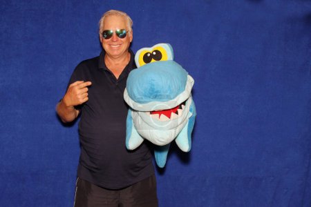 Photo for A man in sunglasses smiles and holds a shark toy as he poses for his picture to be taken while in a Photo Booth at a Party. - Royalty Free Image