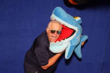 Foto de Un hombre sonríe y ríe mientras posa con un juguete de tiburón para que su foto sea tomada mientras está en una cabina de fotos en una fiesta. - Imagen libre de derechos