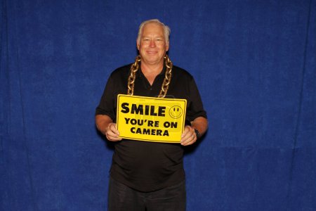 Téléchargez les photos : Souris, tu es filmé. Un homme sourit et tient un sourire que vous êtes sur CAMERA signe en attendant que ses photos soient prises dans un photomaton. - en image libre de droit