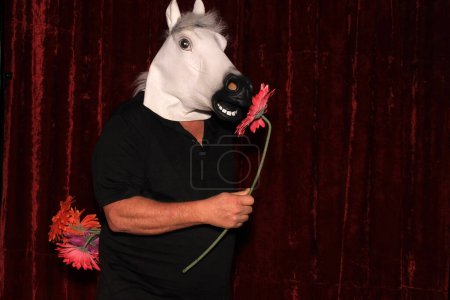 Téléchargez les photos : Un homme portant un masque de tête de cheval en caoutchouc pose et tient des fleurs pendant qu'il se fait prendre en photo dans une cabine photo. - en image libre de droit