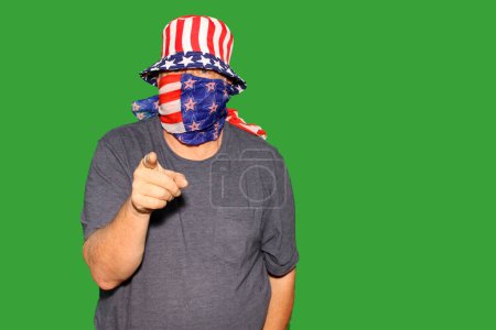 Téléchargez les photos : Écran vert. Chromakey. Un homme porte un chapeau de drapeau américain et le masque de visage pointe vers la caméra tandis qu'en face d'un mur d'écran vert Chromakey aka. Écrans verts sont utilisés pour les effets spéciaux et l'édition - en image libre de droit
