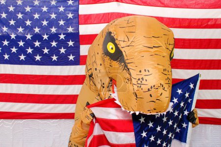 Téléchargez les photos : Homme en costume de dinosaure tient un panneau en carton écrit à la main tout en se tenant devant un drapeau américain dans un photomaton. - en image libre de droit