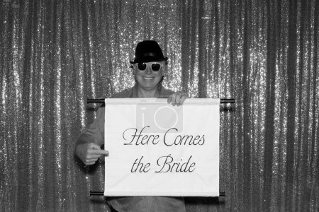Téléchargez les photos : Une photo en noir et blanc d'un homme tient un panneau qui dit ICI COMES THE BRIDE en attendant que ses photos soient prises dans une cabine photo lors d'un mariage ou d'une fête. - en image libre de droit