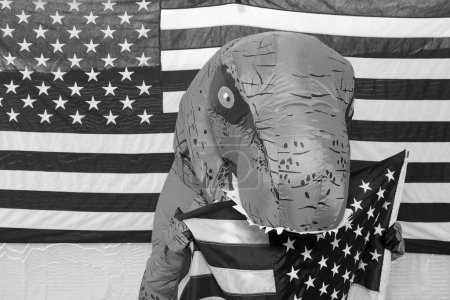 Foto de Hombre con un disfraz de dinosaurio sostiene un letrero de cartón escrito a mano mientras está parado frente a una bandera estadounidense en una cabina de fotos. - Imagen libre de derechos