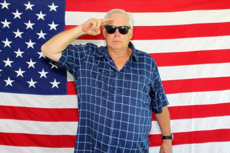 Foto de Un hombre americano orgulloso saluda y posa delante de la bandera americana mientras espera que sus fotos sean tomadas mientras está en una cabina de fotos en una fiesta del 4 de julio. - Imagen libre de derechos