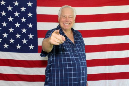 Foto de Un hombre americano orgulloso apunta a la cámara y posa frente a la bandera americana mientras espera a que sus fotos sean tomadas mientras está en una cabina de fotos en una fiesta del 4 de julio. - Imagen libre de derechos