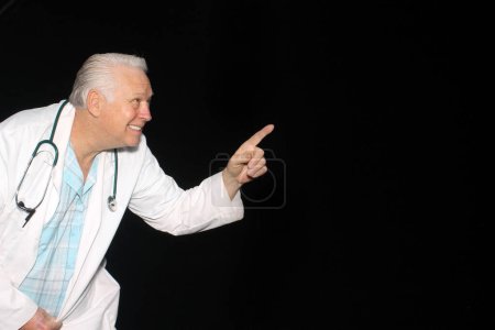 Photo for A friendly and kindly doctor smiles as he points at a space for text while on a black background. Smiling Caucasian male doctor wearing a white medical uniform and stethoscope. - Royalty Free Image
