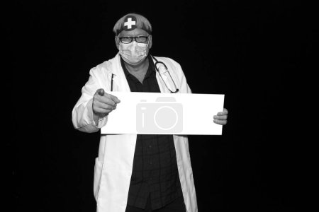 Téléchargez les photos : Médecin. Un docteur maléfique pose alors qu'il se fait photographier dans une cabine photo avec un fond de velours noir. Méfiez-vous de la mauvaise médecine. Le docteur tient un panneau vierge pour la publicité. Médecine. - en image libre de droit