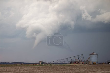 A white cone tornado hangs beneath a storm cloud over rural farmland with farm buildings and equipment in the foreground.