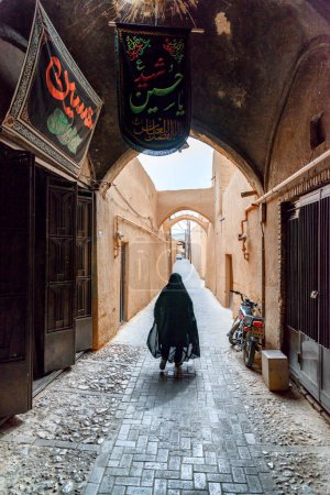 Foto de Yazd, Irán - 26 de octubre de 2018: Mujer iraní vestida de chador negro que camina por la estrecha calle de la histórica ciudad de Yazd. Arquitectura persa única de la antigua ciudad
. - Imagen libre de derechos