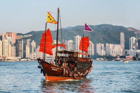 Photo for Hong Kong - October 21, 2017: Amazing view of traditional Chinese wooden sailing ship with red sails in Victoria harbor. High-rise residential buildings are visible at Hong Kong Island. - Royalty Free Image