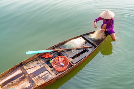 Top view of Vietnamese woman in traditional bamboo hat on wooden boat checking her fishing net on the Thu Bon River at Hoi An Ancient Town, Vietnam. Hoian is a popular tourist destination of Asia.