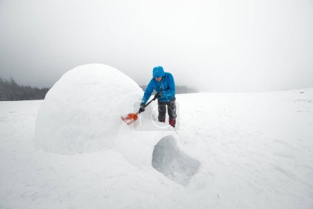 Téléchargez les photos : Homme en veste bleue construisant igloo dans la haute montagne. Fantastique scène d'hiver - en image libre de droit