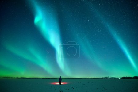 Foto de Turista con linterna roja en el campo nevado contra el telón de fondo del increíble cielo estrellado con Aurora boreal. Increíble paisaje nocturno. Luces boreales en campo de invierno - Imagen libre de derechos