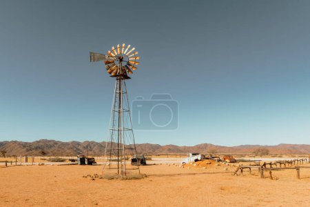 Téléchargez les photos : Abandoned derelict old car graveyard and old rusty wind turbine in the sandy desert with a blue sky on the background - en image libre de droit