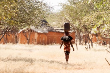 Téléchargez les photos : OPUWO, OTJIWARONGO REGION NAMIBIA - MAY 4, 2021: Young unidentified Himba woman with the typical dresses and water canister on head in Himba living museum during a appearance in front of tourists - en image libre de droit