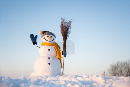 Photo for Funny snowman in knitted hat and yellow scalf with broom on snowy field. Blue sky on background - Royalty Free Image