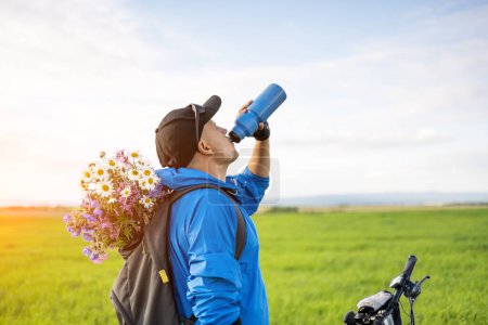 Téléchargez les photos : Homme avec vélo eau potable dans le champ de printemps. Bouquet de fleurs sauvages dans un sac à dos de motard - en image libre de droit