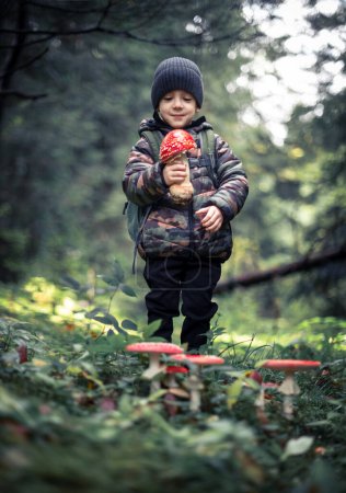 Téléchargez les photos : Petit enfant avec Fly agaric Amanita muscari, beau champignon toxique hallucinogène à tête rouge dans la forêt d'automne. Enfance avec concept amoureux de la nature - en image libre de droit