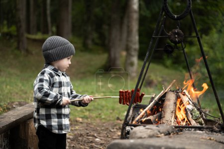 Foto de Chico fríe salchichas en fogata en el bosque de verano. Infancia con concepto de amor a la naturaleza - Imagen libre de derechos