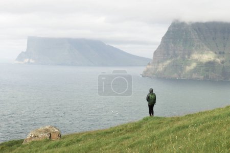 Foto de El turista solitario cerca de un pequeño lago mira las islas de niebla en el océano Atlántico desde la isla Kalsoy, Islas Feroe, Dinamarca. Paisaje fotografía - Imagen libre de derechos