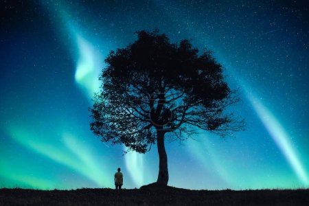 Foto de Solitario humano bajo un viejo árbol en el campo nocturno contra el telón de fondo del increíble cielo con Aurora Boreal Luz del Norte. Paisaje fotografía - Imagen libre de derechos