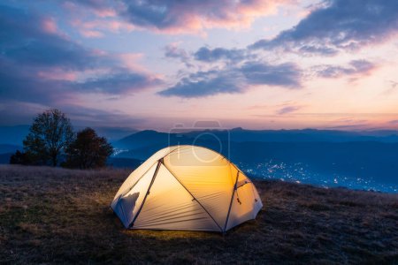 Téléchargez les photos : Tente touristique éclairée de l'intérieur par une lumière chaude sur fond de paysage montagneux incroyable au coucher du soleil - en image libre de droit