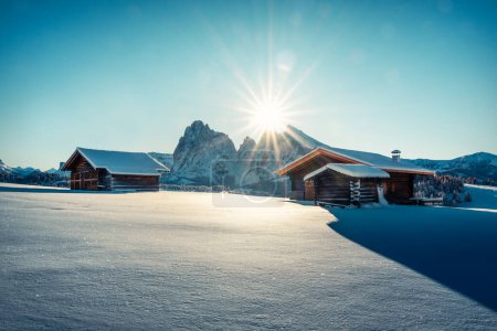 Photo for Small wooden log cabins on snowy meadow Alpe di Siusi on blue sky background with sun. Seiser Alm, Dolomites, Italy. Landscape photography - Royalty Free Image