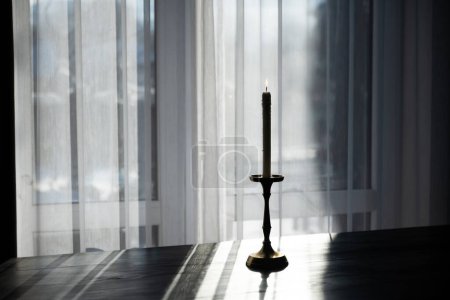 Téléchargez les photos : Bougie d'éclairage dans un chandelier en laiton sur la table sur le fond d'une fenêtre avec tulle - en image libre de droit