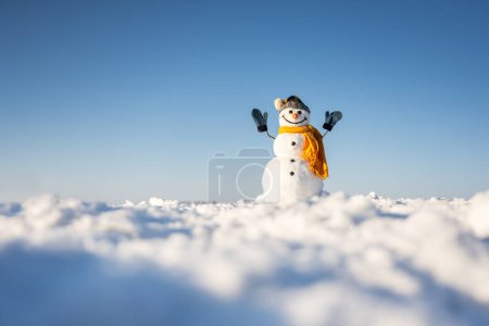 Foto de Muñeco de nieve divertido en sombrero de punto y cuero cabelludo amarillo con las manos en el campo cubierto de nieve. Cielo azul sobre fondo. Concepto vacaciones de invierno - Imagen libre de derechos