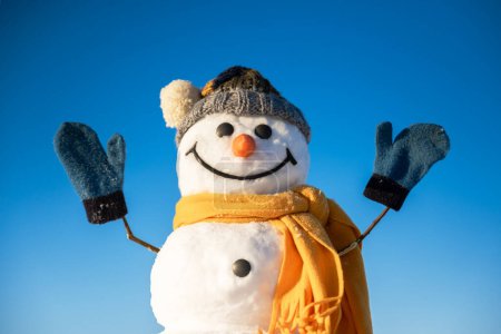 Photo for Funny snowman in knitted hat and yellow scalf with hands up on blue sky background. Winter holidays concept - Royalty Free Image