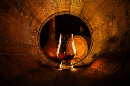 Foto de Un vaso de whisky escocés en barrica de roble viejo. Cobre alambico y pequeño barril sobre fondo. Concepto tradicional de destilería de alcohol - Imagen libre de derechos
