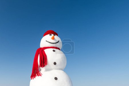 Téléchargez les photos : Dans un paysage enneigé, un bonhomme de neige joyeux porte un chapeau rouge élégant et une écharpe rouge assortie, le tout sous un ciel bleu vif. Carte postale de Noël - en image libre de droit