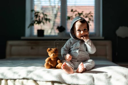 Téléchargez les photos : Petit garçon en pyjama gris assis sur le lit avec son ours en peluche préféré. Concept d'enfance heureuse - en image libre de droit
