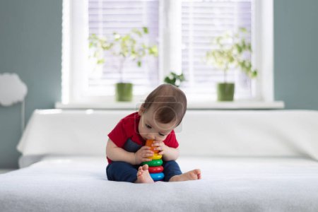 Téléchargez les photos : Petit garçon pieds nus en t-shirt rouge mangeant son jouet préféré pyramide colorée. Concept d'enfance heureuse - en image libre de droit