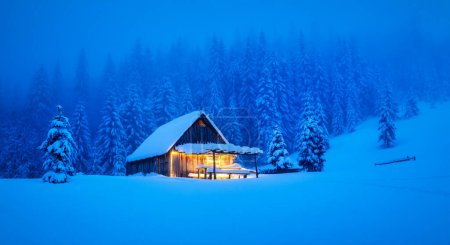 Téléchargez les photos : Une scène hivernale au cœur des bois avec un chalet en bois solitaire et des pins enneigés sur une clairière de montagne. Carte postale de Noël. Forêt de montagnes enneigées - en image libre de droit