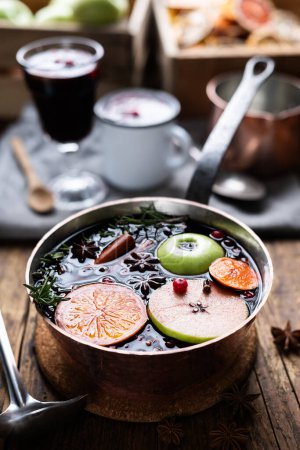 Photo for Mulled wine with slices of orange, apple, cinnamon, star anise, rosemary and cranberries in copper pot on kitchen table. Food photography - Royalty Free Image