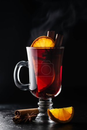 Téléchargez les photos : Vin chaud chaud dans un verre avec une tranche d'orange fraîche et des bâtonnets de cannelle sur un fond noir. Photographie gastronomique - en image libre de droit