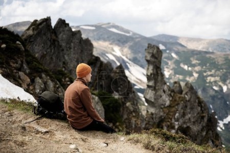 Foto de Un turista con mochila se sienta en el borde de un pico de montaña. Montañas nevadas en el fondo. Paisaje fotografía - Imagen libre de derechos