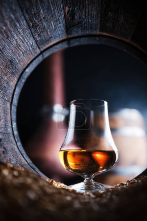 Foto de Un vaso de whisky en barrica de roble viejo. Destilador de cobre sobre fondo. Concepto tradicional de destilería de alcohol - Imagen libre de derechos