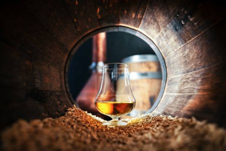 Téléchargez les photos : Un verre de whisky bourbon dans un vieux fût de chêne. Cuivre alambique et fût de chêne sur fond. Concept traditionnel de distillerie d'alcool - en image libre de droit