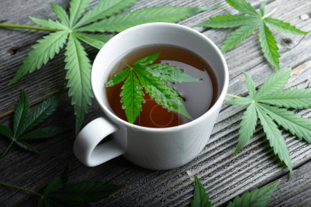 Photo for Cannabis tea with green marijuana leaves in white cup on wooden table. Medical herbal tea with cannabis close up - Royalty Free Image